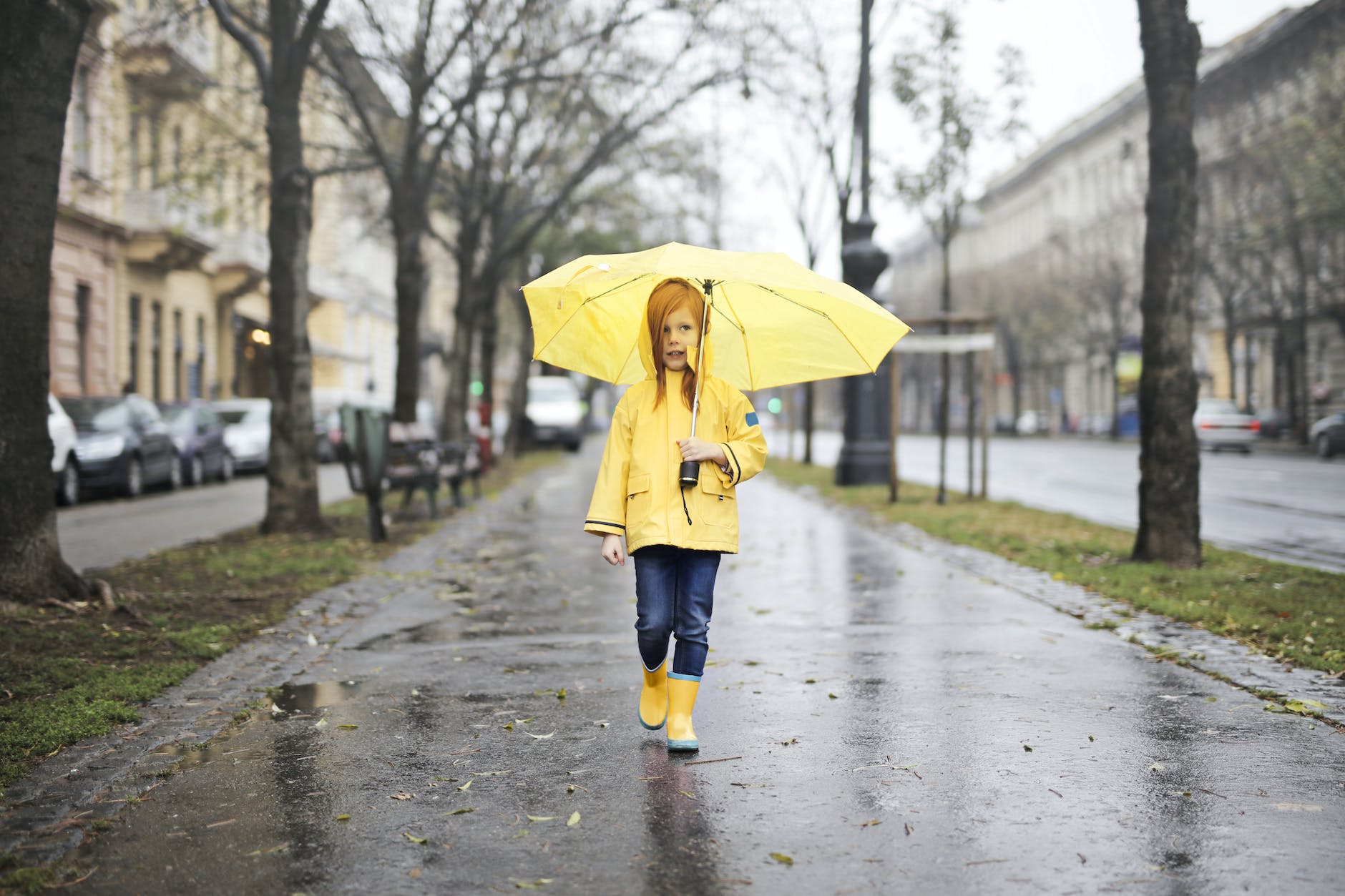 girl in yellow raincoat and yellow boots holding yellow umbrella walking on gray concrete side walk