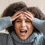Expressive African American woman with curly brown hair touching head and yelling against light background